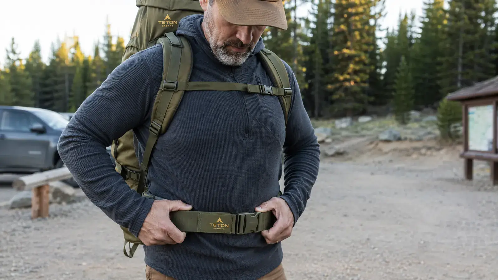 Camper tightening the hip belt of an internal frame backpack at a trailhead before a hike, trees in background