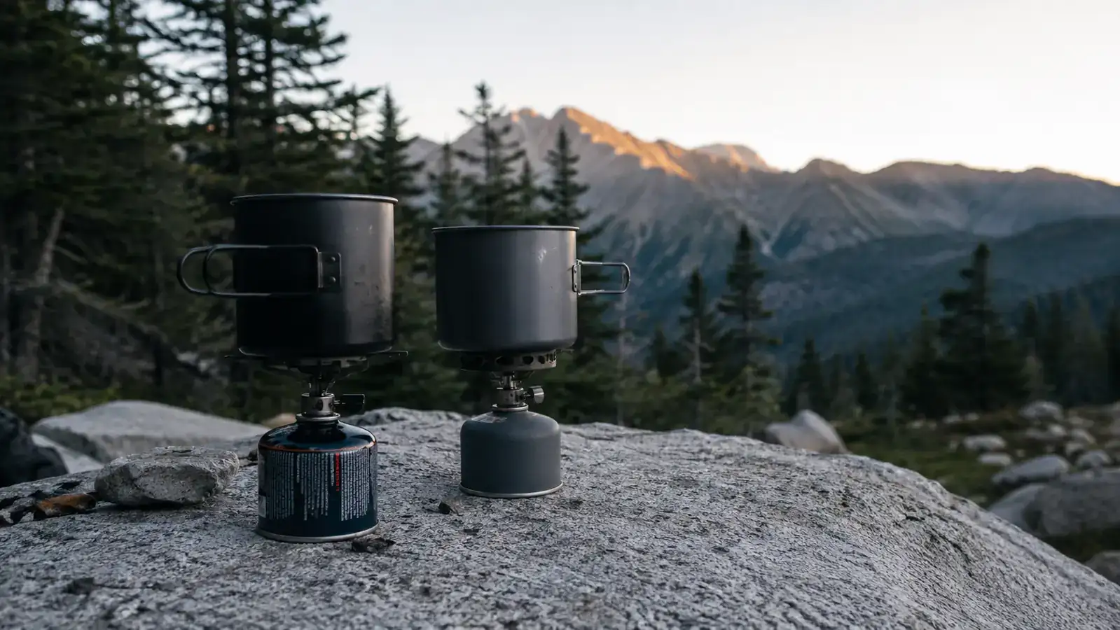 Two backpacking stoves side by side on a granite boulder at a mountain campsite, morning light, one compact canister-mount stove and one integrated system