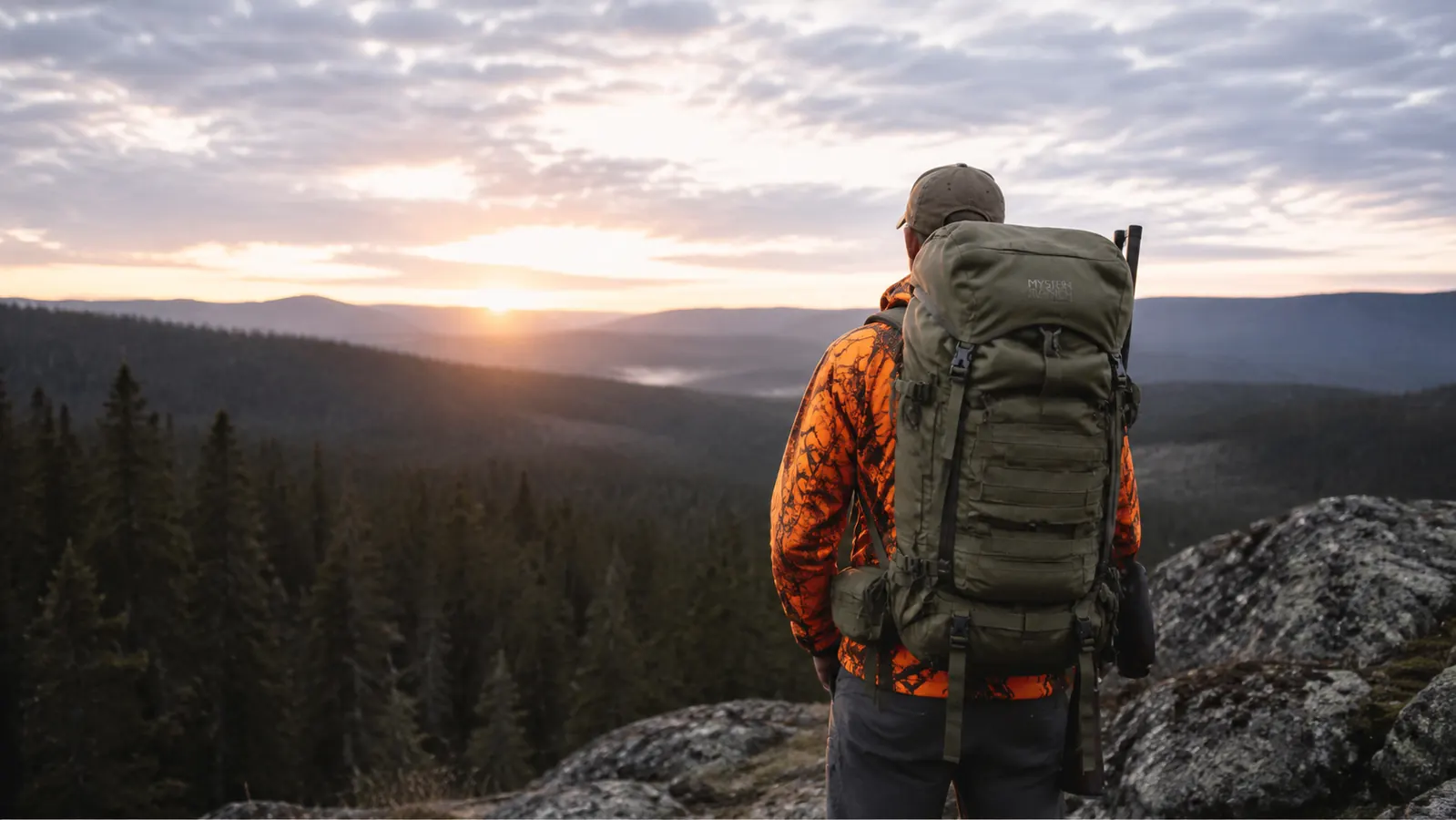 Hunter with large internal frame backpack on a mountain ridgeline at dawn, conifer forest below