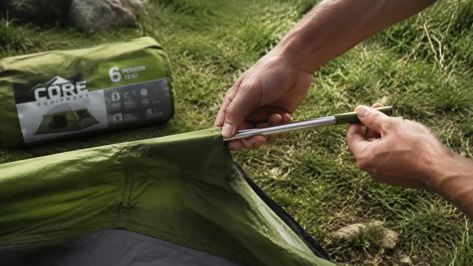 Camper threading a pole through the dome tent sleeve during setup at a grassy campsite