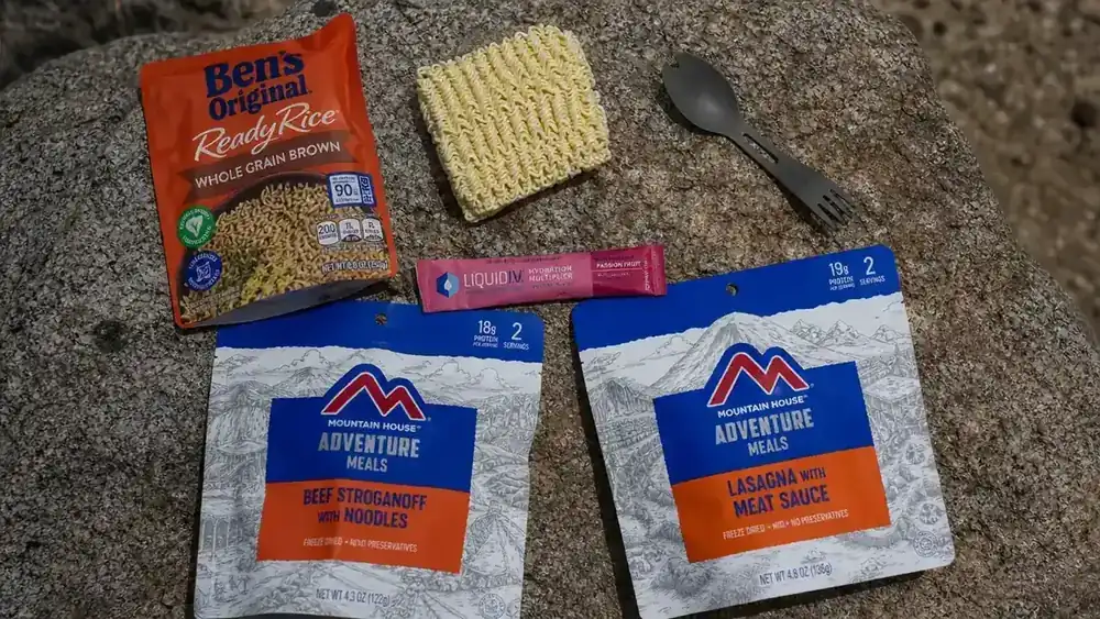 Trail food ingredients laid out on a rock: instant rice packets, freeze-dried meal pouches, ramen, and an electrolyte packet