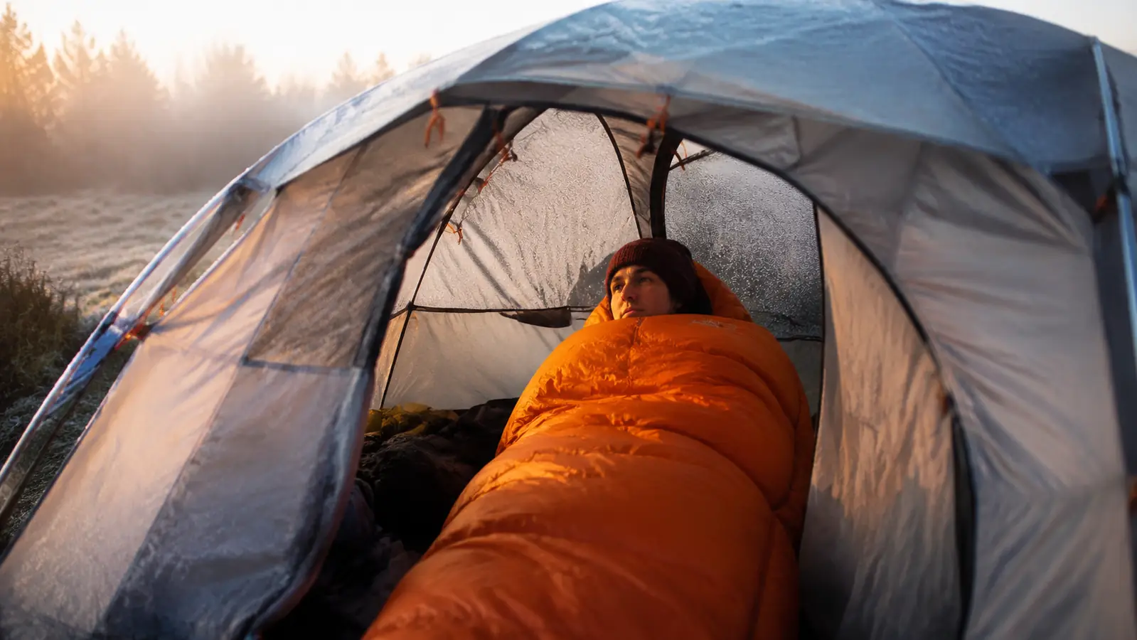 Camper in a mummy sleeping bag inside a dome tent on a cold autumn morning, frost visible on the rainfly