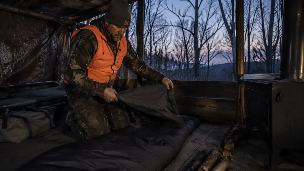 Hunter unrolling a mummy sleeping bag at a deer camp in a late-autumn forest at dusk
