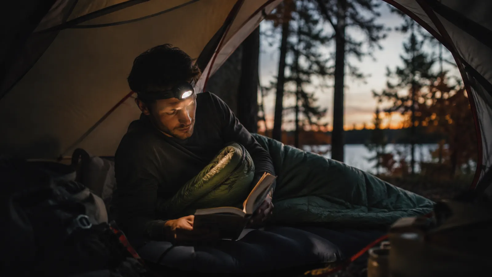 Campsite at dusk with a sleeping bag laid out on a sleeping pad inside a tent, headlamp glowing nearby